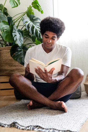 Young african american man sitting cross-legged on the floor reading a book. Vertical image. At home concept.